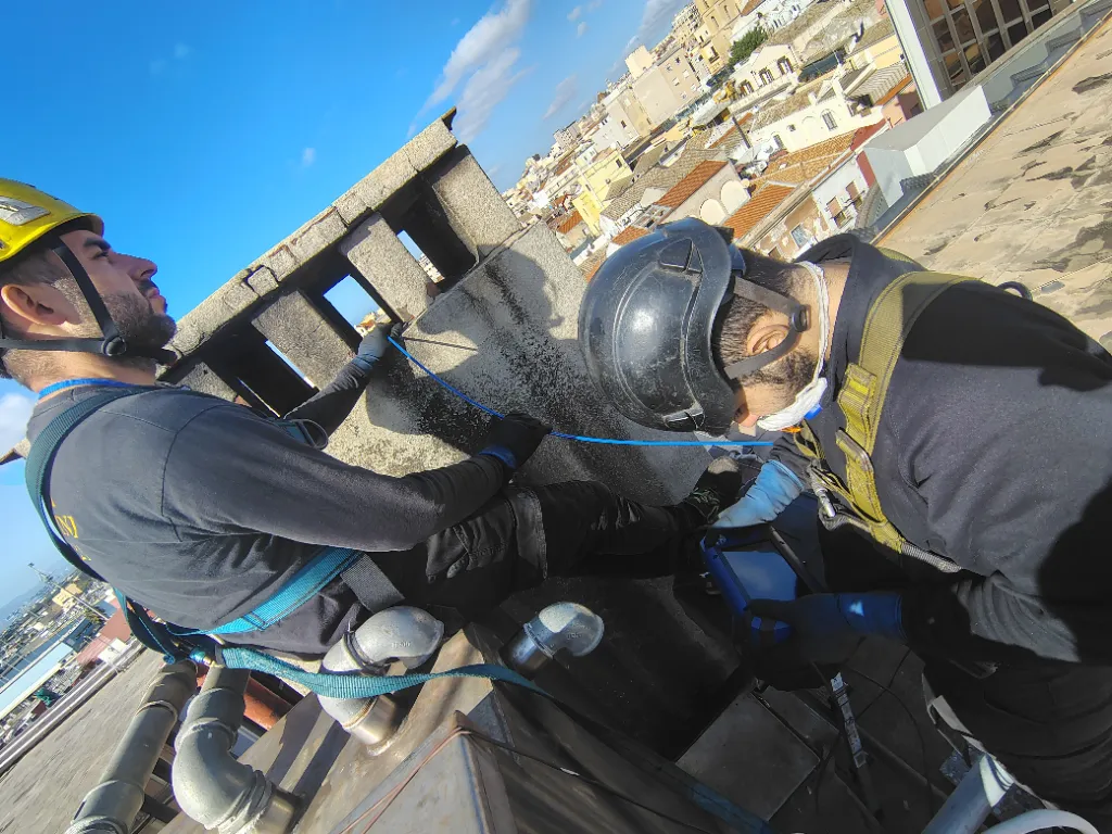 Due spazzacamini SardaCAMINI con casco e imbracatura al lavoro su un tetto di Cagliari