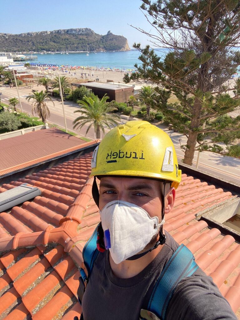 Spazzacamino con casco, mascherina e imbracatura sul tetto, con il Poetto e la Sella del Diavolo sullo sfondo