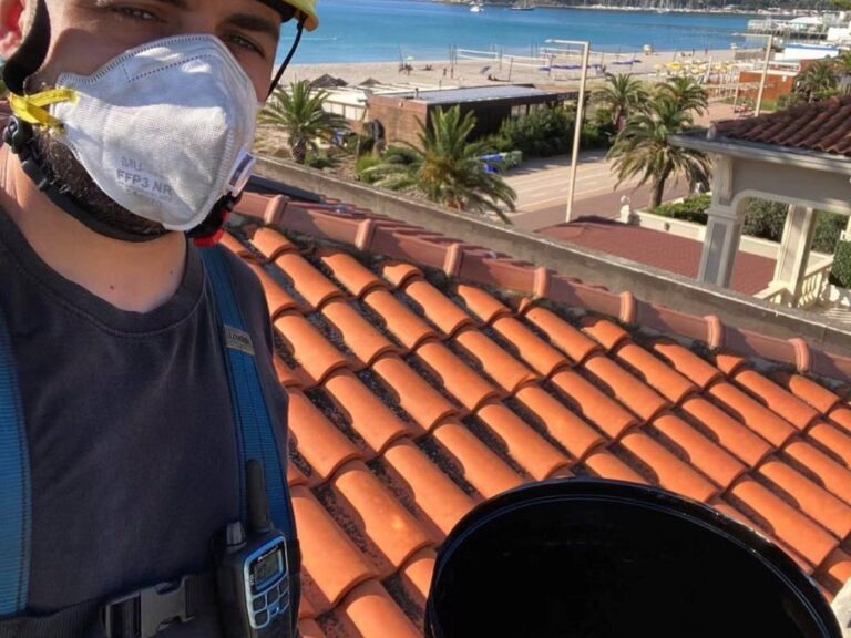 Spazzacamino con casco, mascherina e imbracatura sul tetto accanto alla canna fumaria in acciaio
