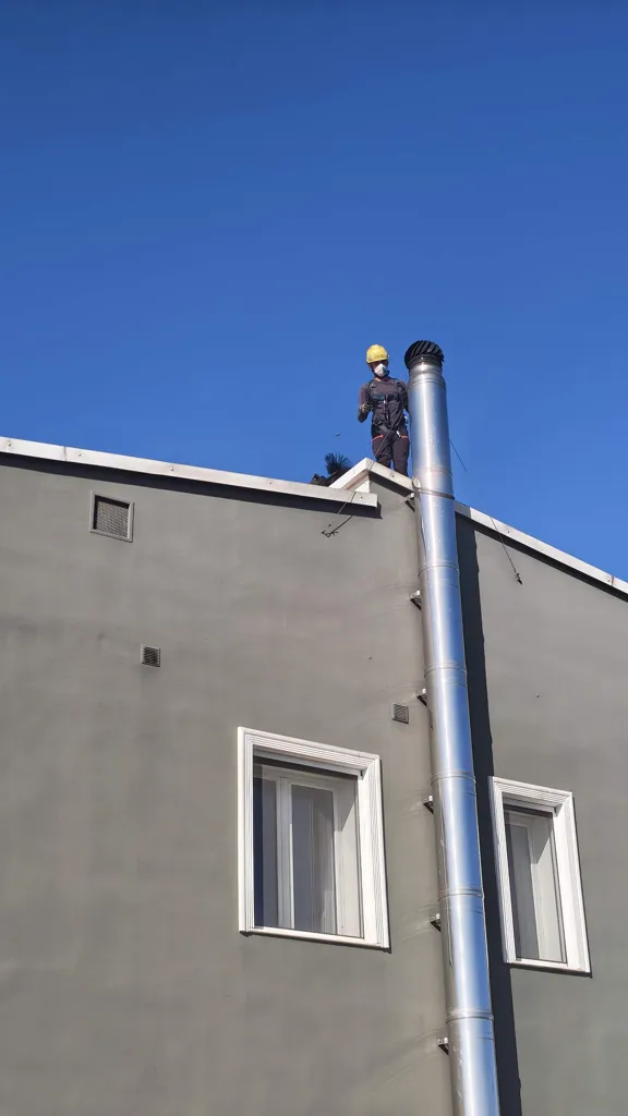 Spazzacamino con casco, mascherina e imbracatura sul tetto accanto alla canna fumaria in acciaio